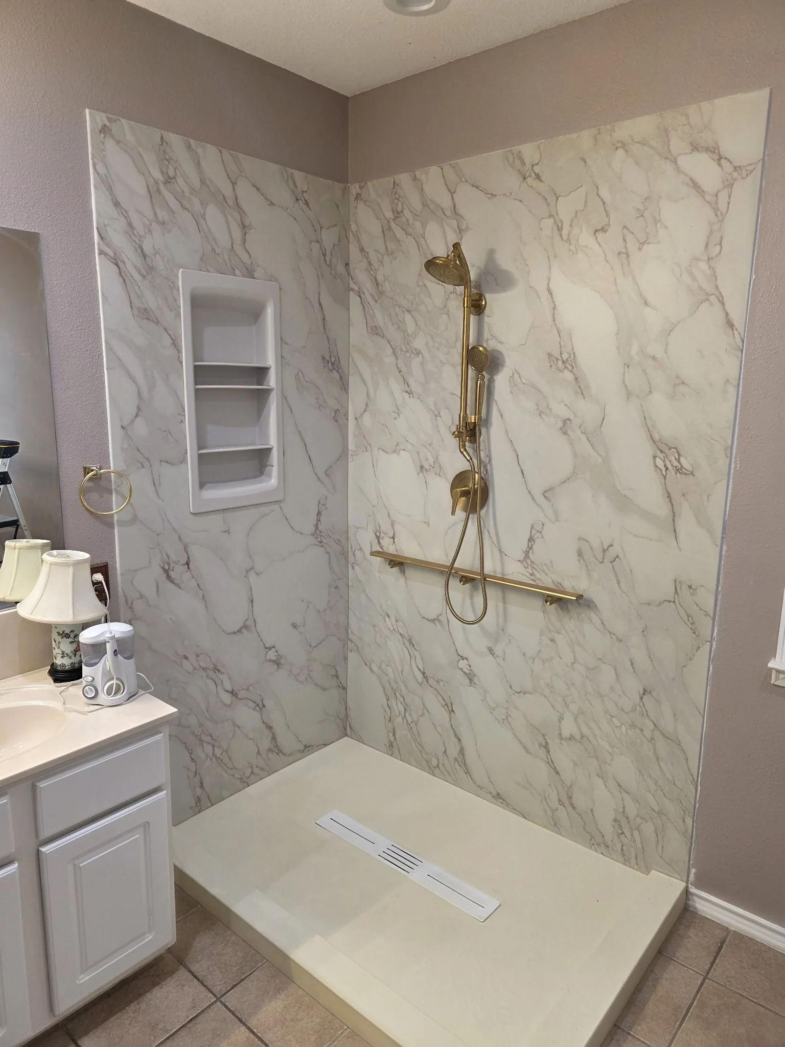 Finished walk-in shower with white marble-pattern walls, gold showerhead and handheld sprayer, gold grab bar, recessed shelves and a low-profile pan with linear drain