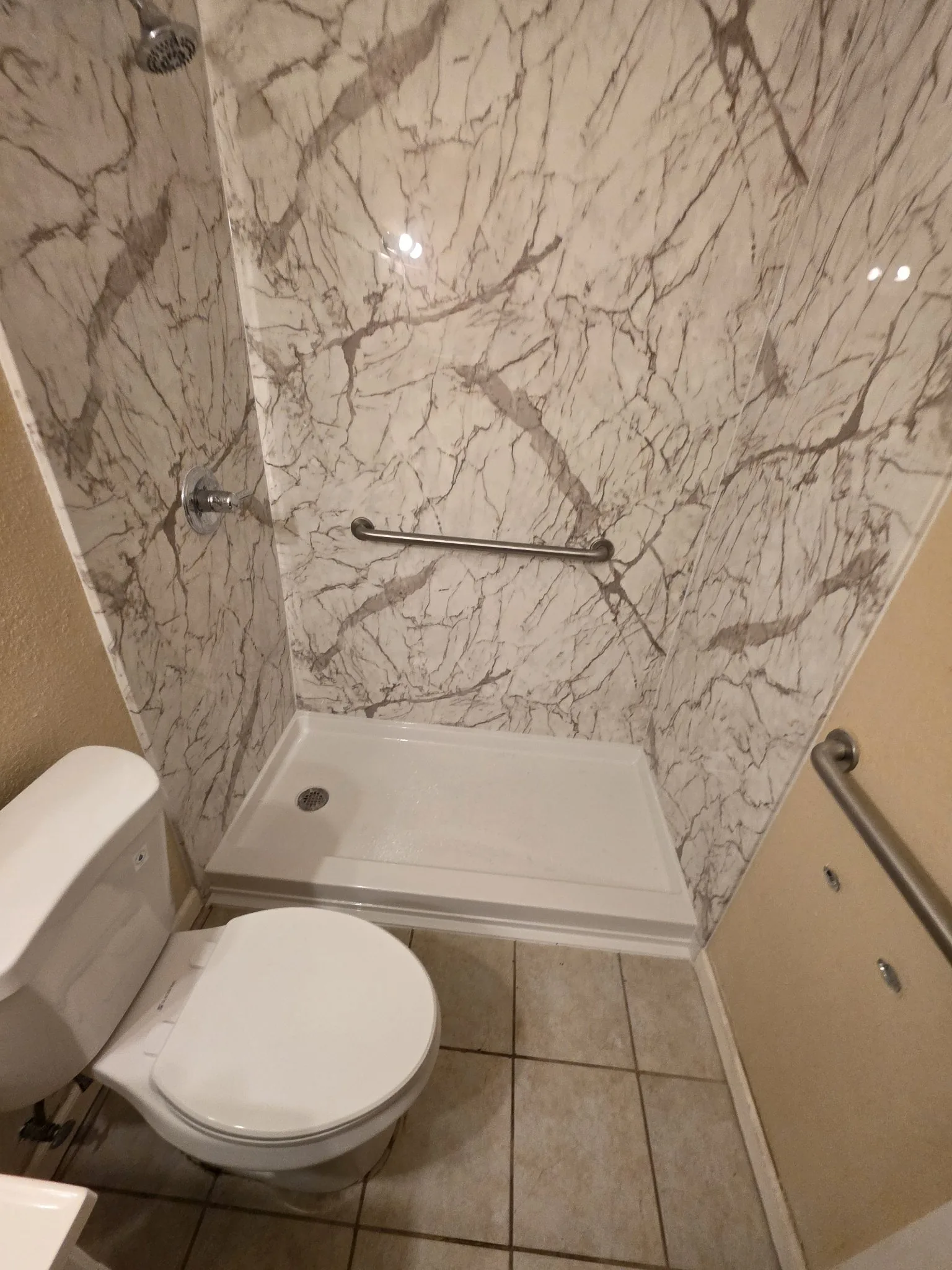 Finished white marble-pattern shower with chrome showerhead and controls, horizontal grab bar and white low-profile pan beside a toilet