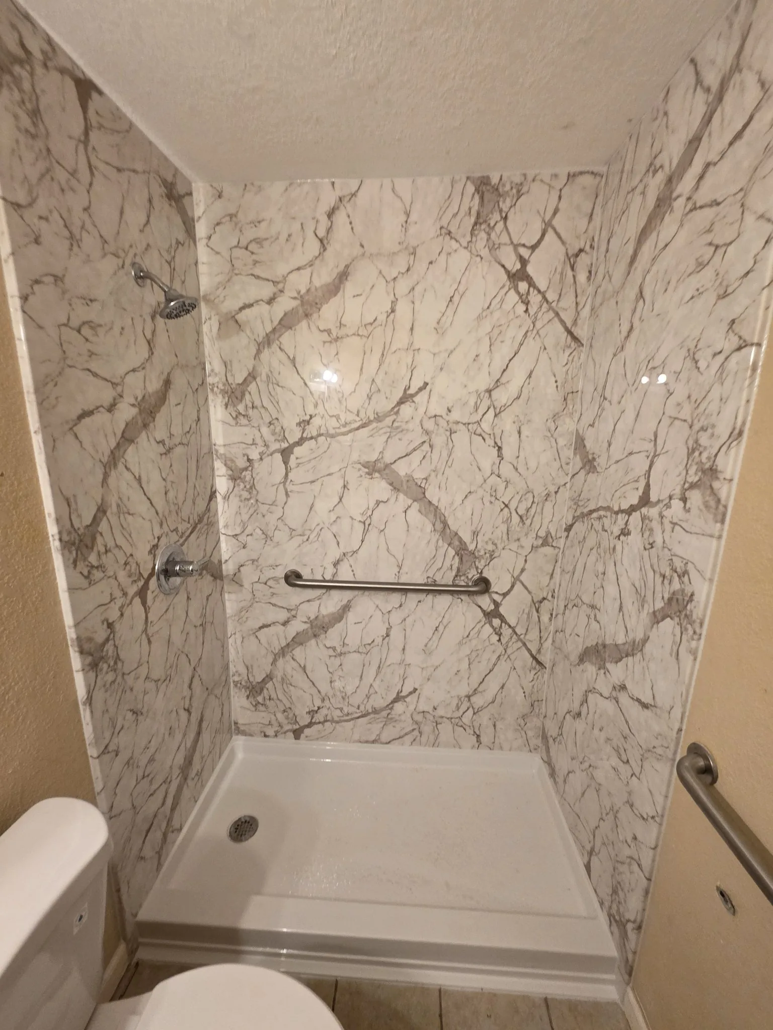 Closer view of a finished white marble-pattern shower with chrome showerhead and controls, horizontal grab bar and white low-profile pan