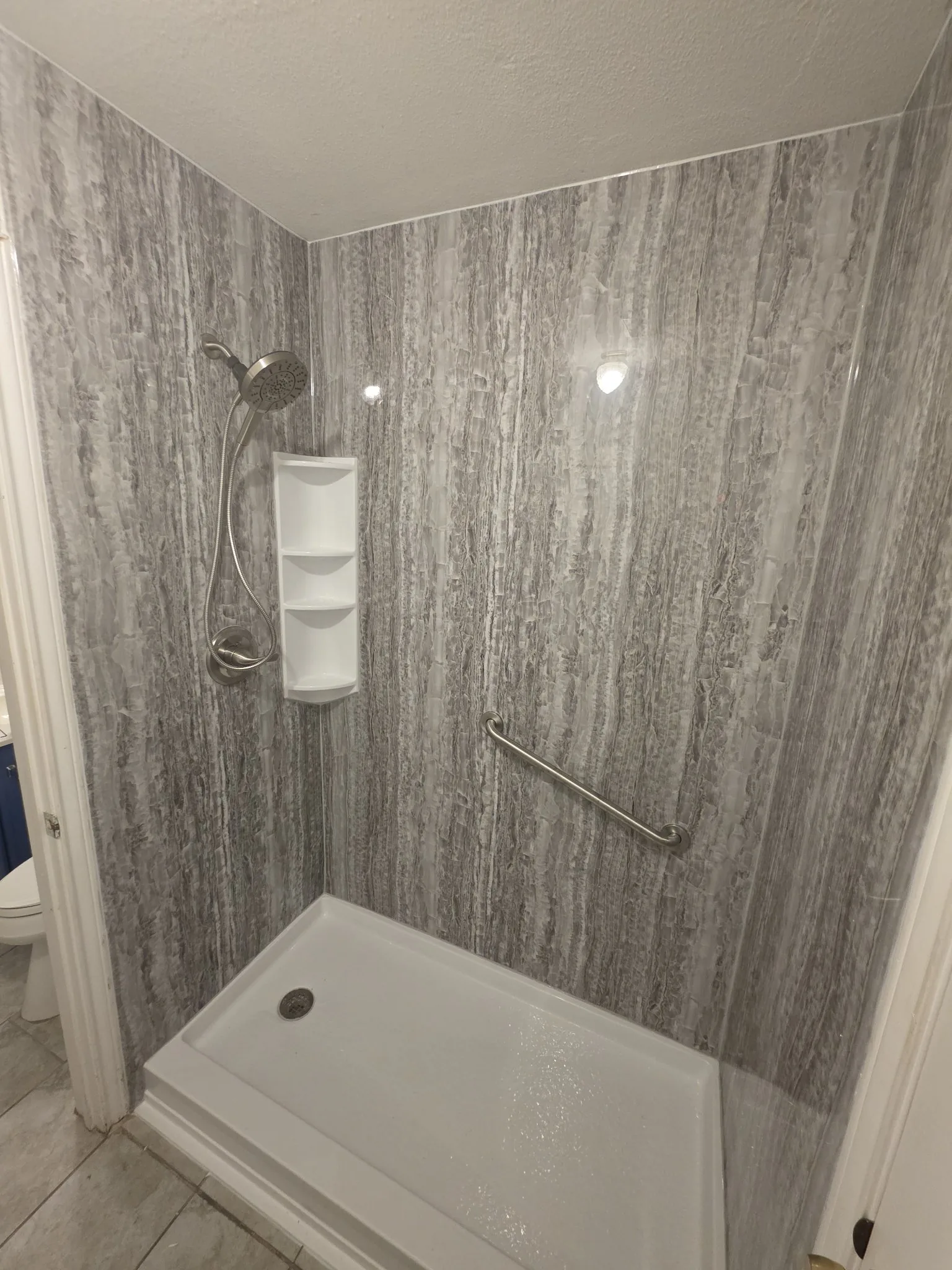 Finished shower with grey streaked walls, brushed-nickel handheld shower, white corner caddy, angled grab bar and white shower pan