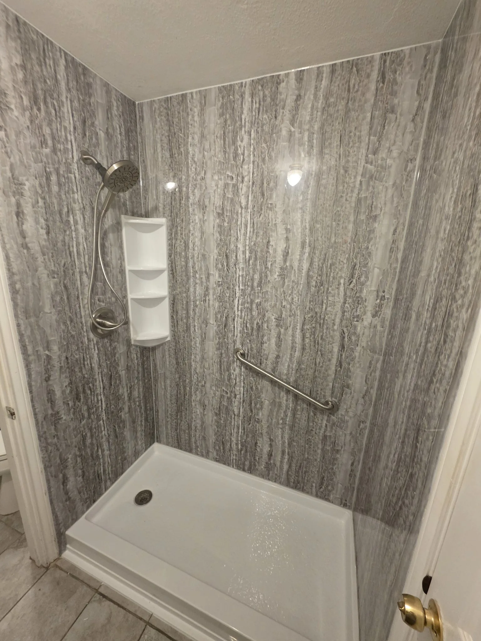 Angled view of a finished grey streaked shower with brushed-nickel handheld shower, white corner caddy, angled grab bar and white pan