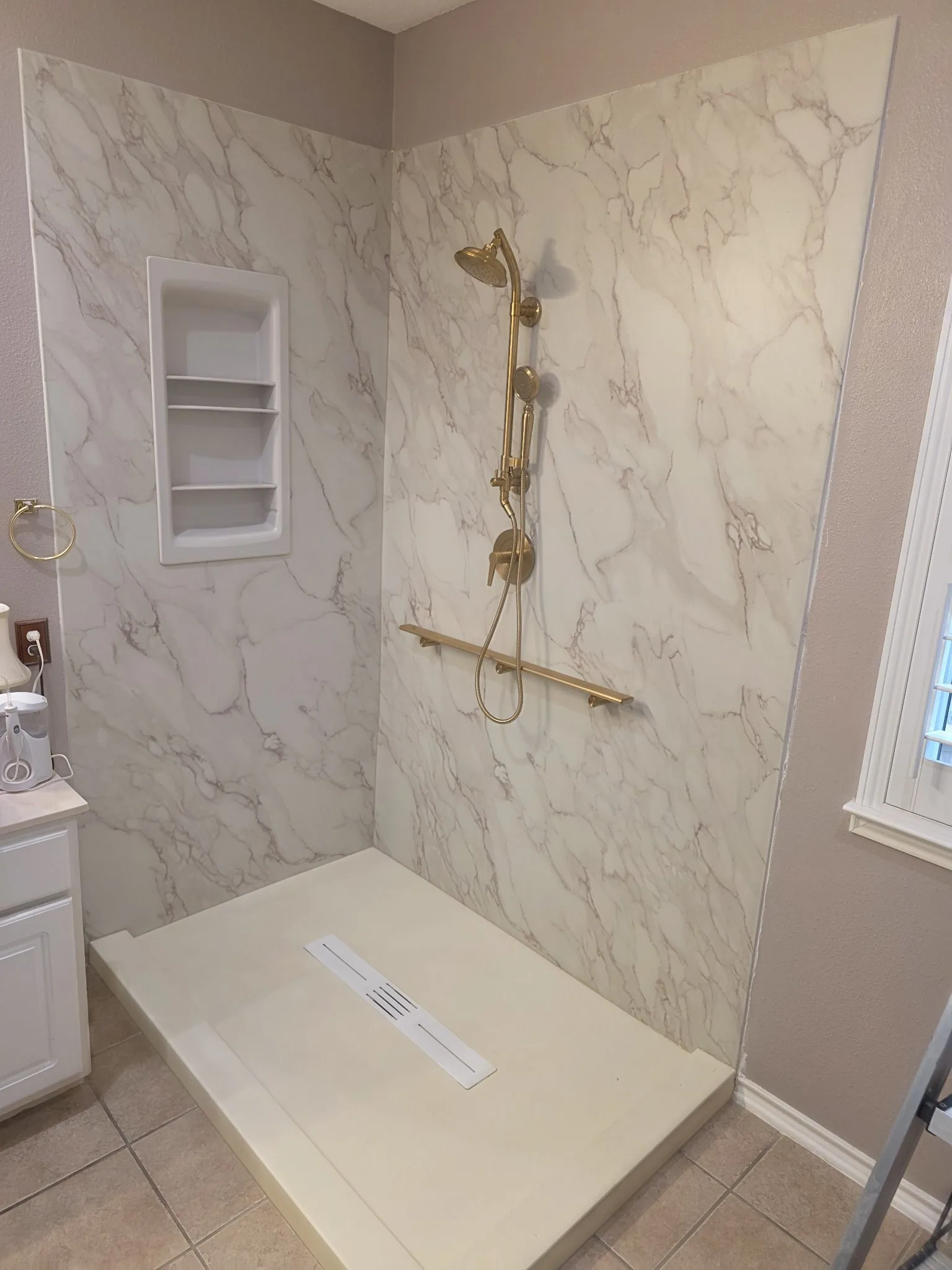 Angled view of a finished white marble-pattern walk-in shower with gold shower fixtures, handheld sprayer, grab bar, recessed shelves and linear drain