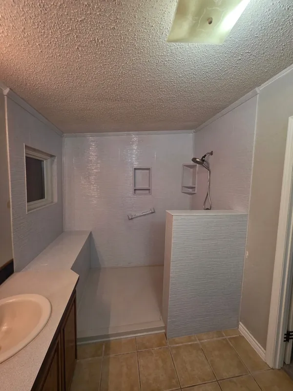 Before photo: dated mobile-home walk-in shower with worn fiberglass walls, popcorn ceiling and original tile floor — ready for a full transformation