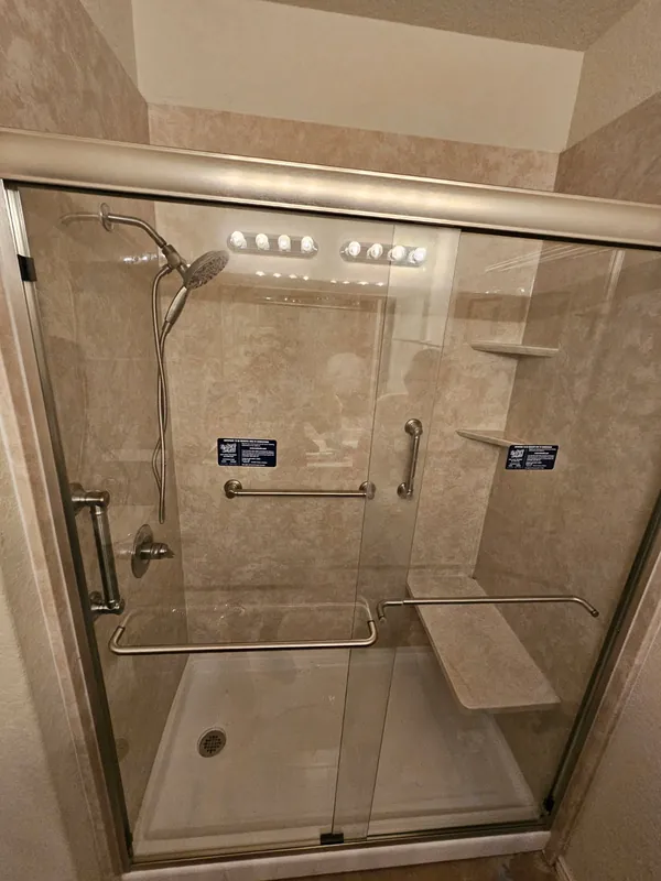 Beige solid-surface walk-in shower with built-in granite bench, multiple grab bars and corner shelves — full aging-in-place setup
