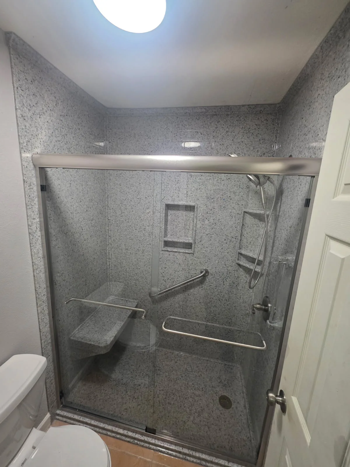Gray granite-pattern walk-in shower with built-in bench, dual brushed-nickel grab bars, recessed niches and bypass glass doors — ADA-friendly bathroom remodel in San Antonio
