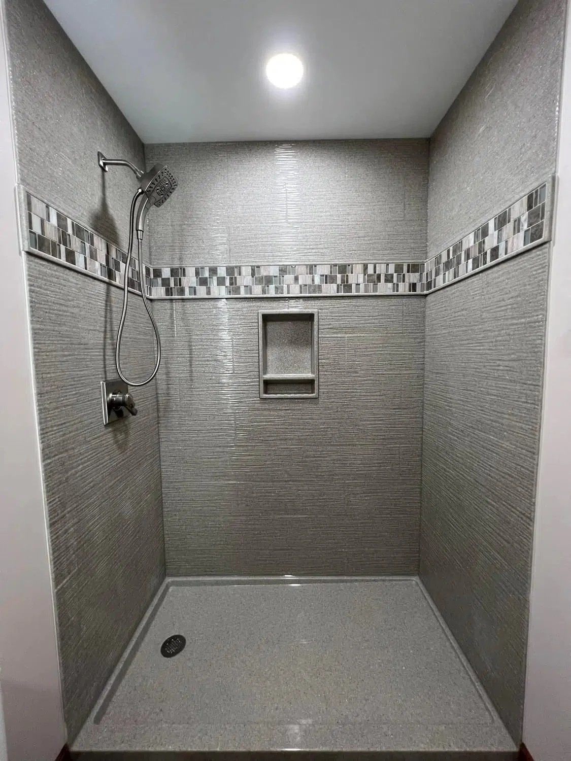 Gray textured solid-surface shower with mosaic glass tile accent band, recessed niche and matching shower pan