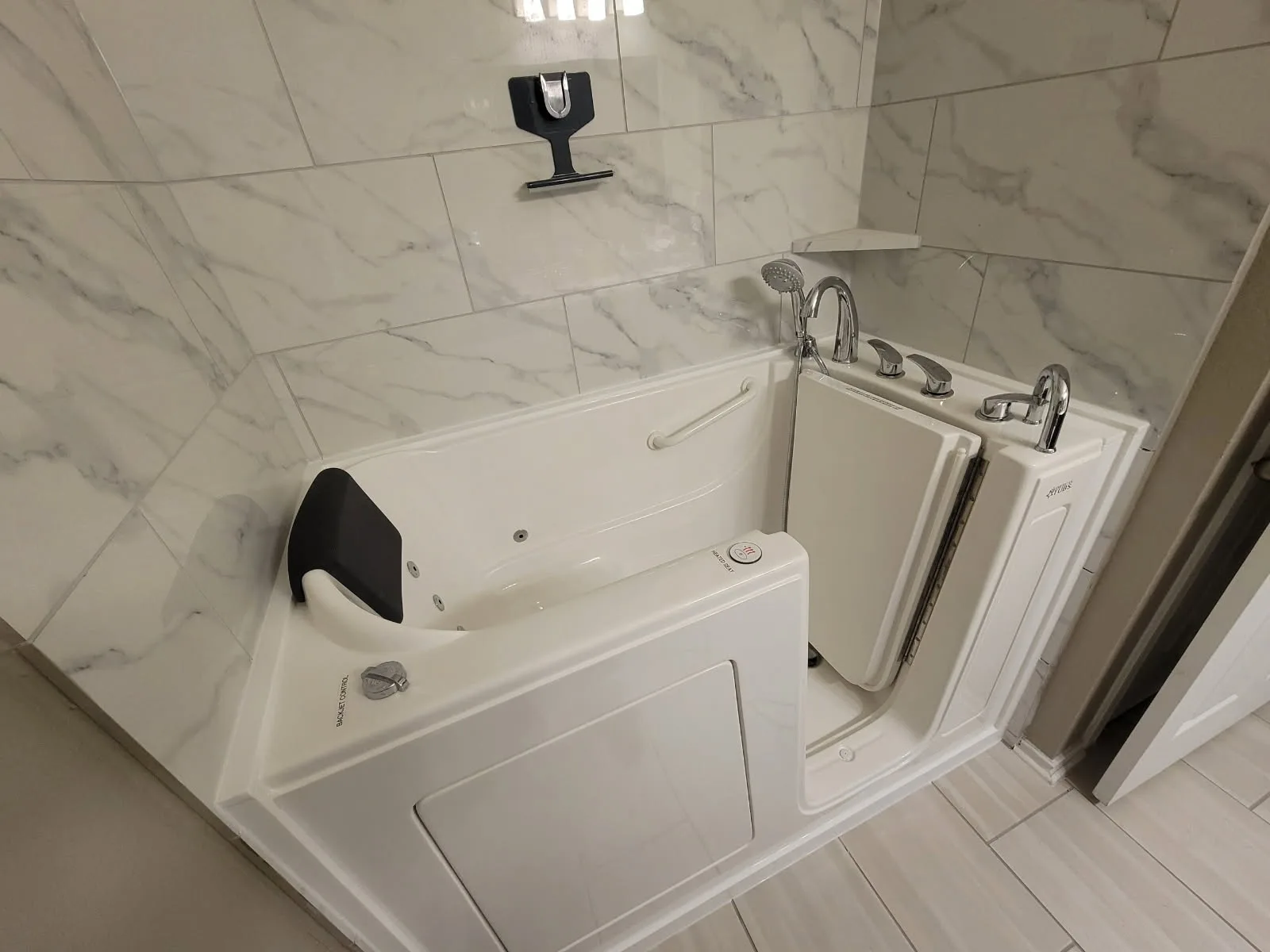 Jetted walk-in tub with hydrotherapy jets, padded headrest and inward-swing door open against a marble-pattern tile surround — accessible bathing remodel by Out This World Services