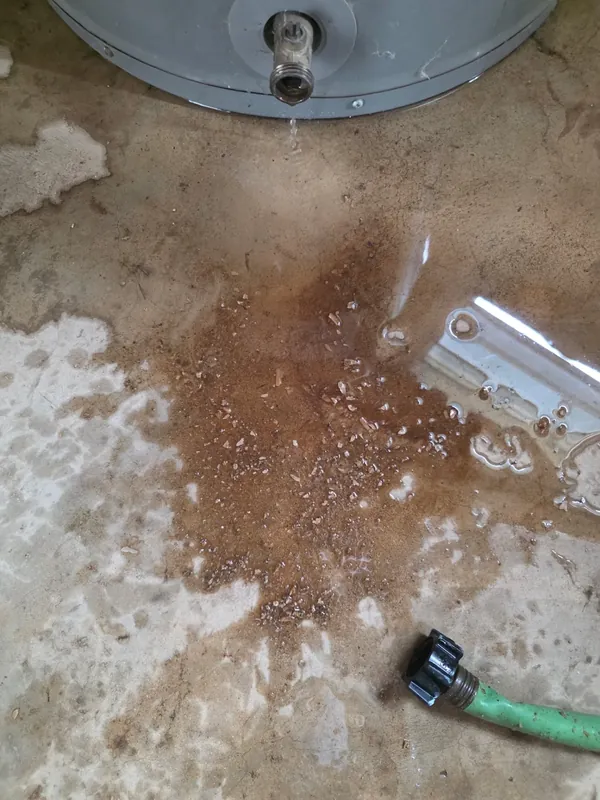 Leaking water heater draining rust-colored sediment onto a garage floor — emergency replacement call in San Antonio
