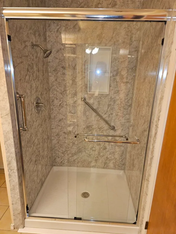 Tan marble-pattern walk-in shower with frameless glass door, chrome grab bars, recessed niche and low-profile shower pan
