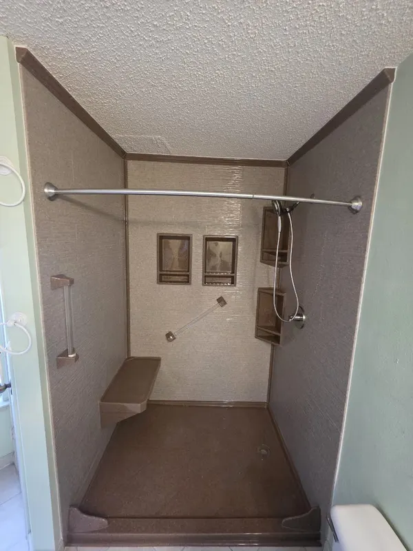 Tan textured solid-surface walk-in shower with built-in bench, ADA grab bar, dual recessed niches and curved chrome rod — aging-in-place remodel