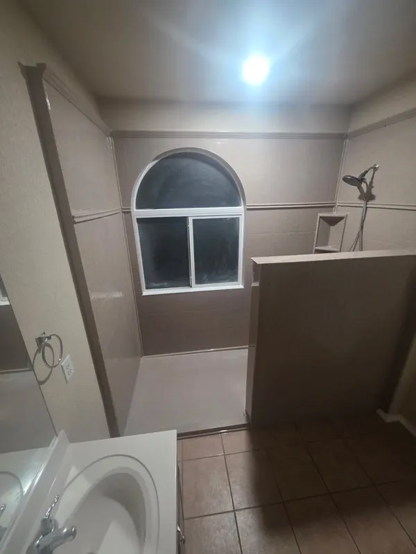 Beige-tile walk-in shower with arched accent window, tiled half-wall divider and handheld sprayer installed in a San Antonio home