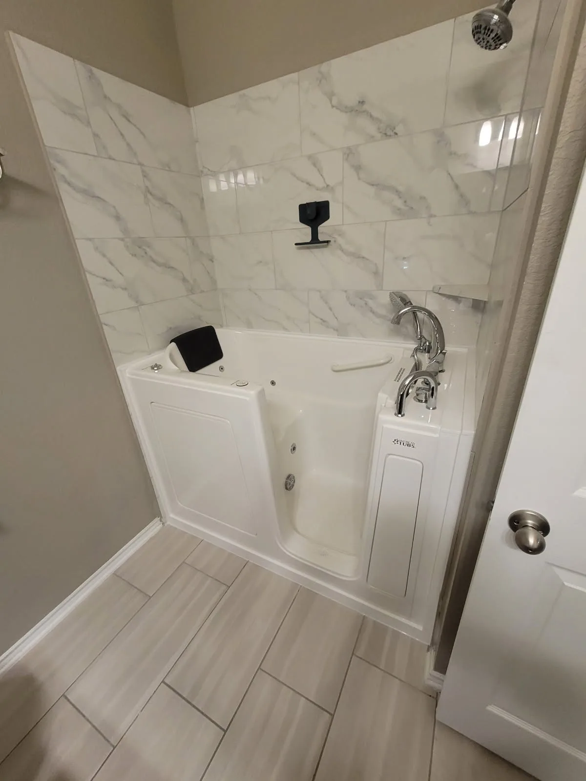 Finished white walk-in tub with chrome handheld shower wand and grab bar, set into a fresh marble-pattern tile alcove — aging-in-place install in a San Antonio bathroom