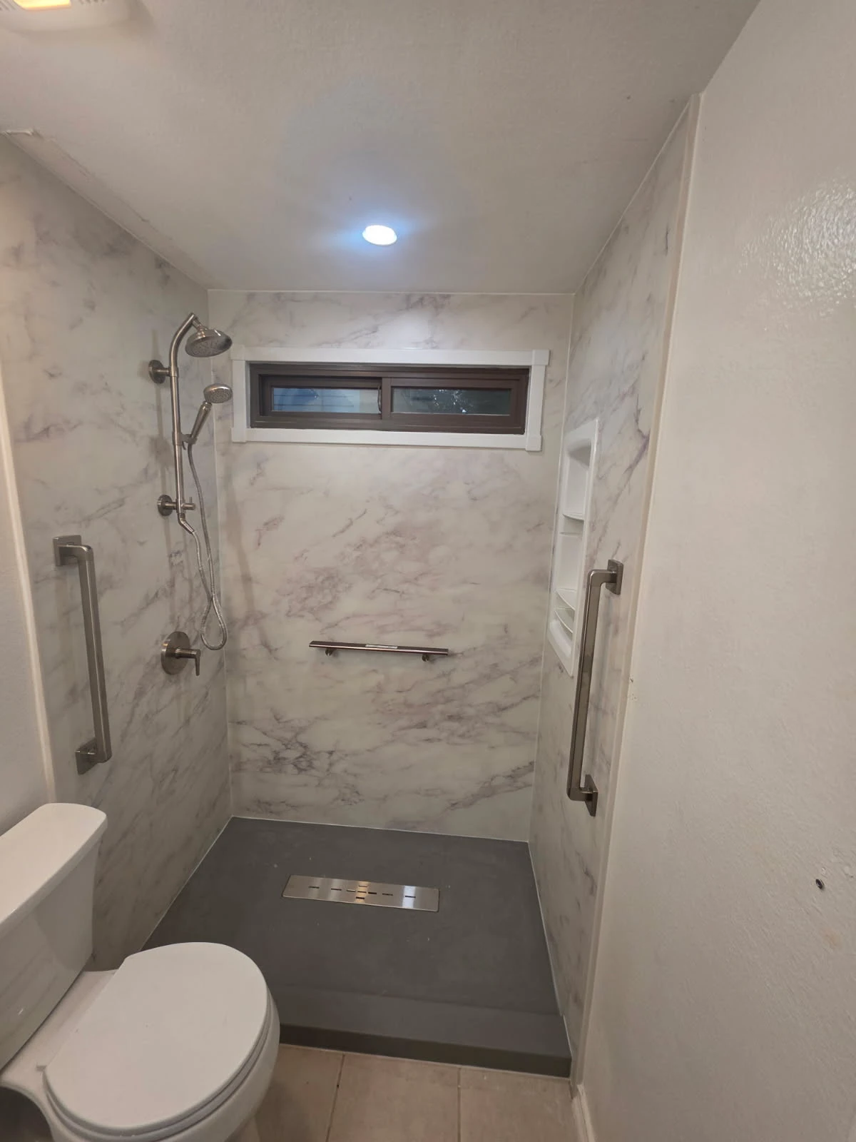 Curbless white marble-pattern walk-in shower with linear drain, brushed-nickel rain shower, dual grab bars and recessed corner shelves for safe aging-in-place bathing