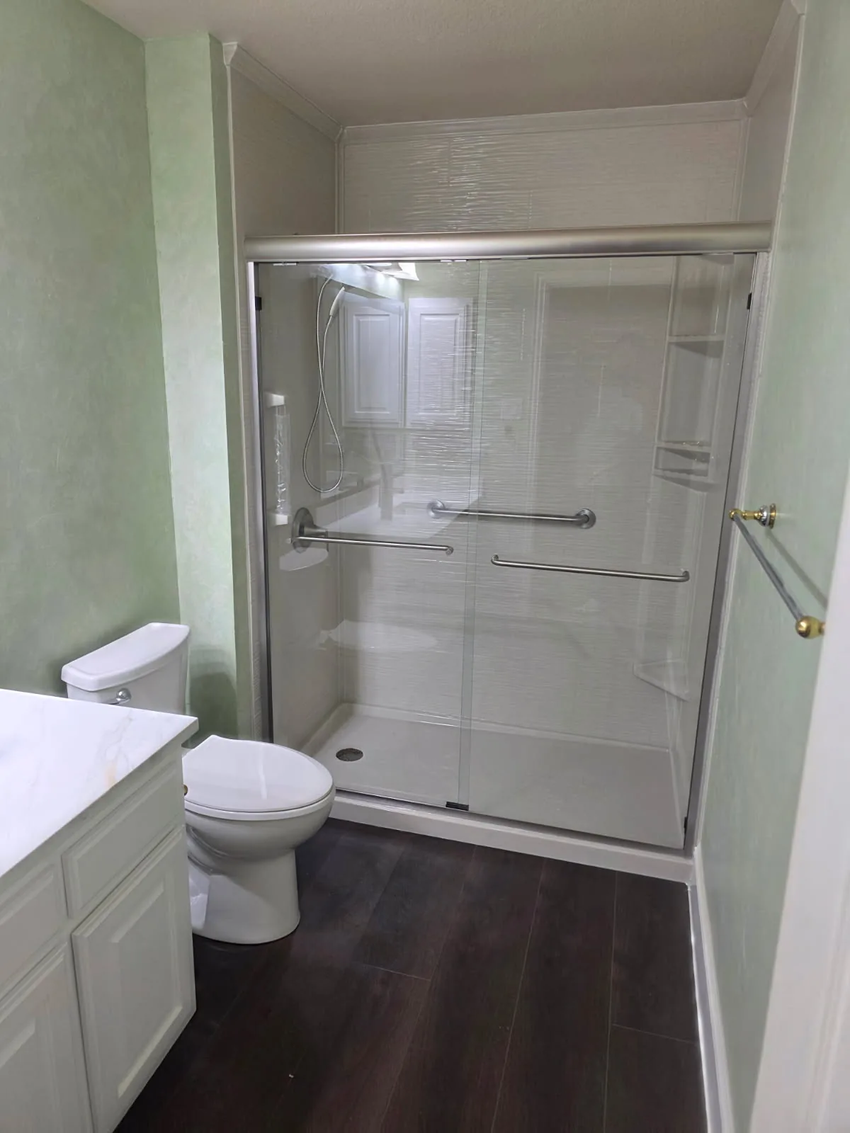 Finished white solid-surface walk-in shower with brushed-nickel bypass glass doors, dual grab bars and low-profile pan installed over dark wood-look flooring — tub-to-shower conversion by Out This World Services