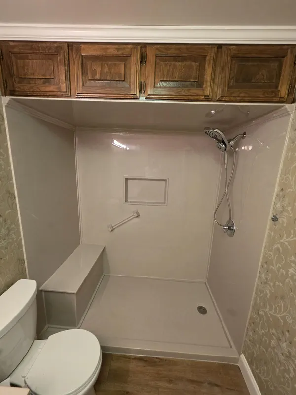 White solid-surface walk-in shower with built-in bench, recessed niche and chrome handheld installed under existing wood cabinets — clean tub-to-shower conversion